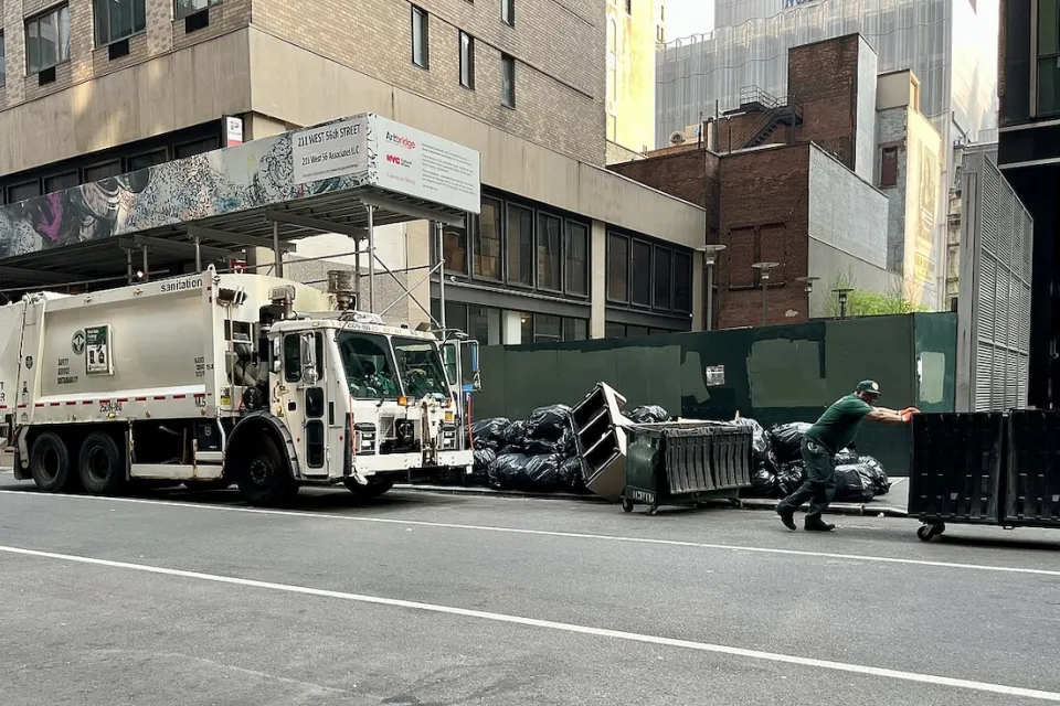 White waste management sanitation garbage truck and worker pushing wheeled green dumpster on street with piles of trash bags.